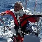Nodas at the helm in a red sailing jacket, in a strong breeze.
