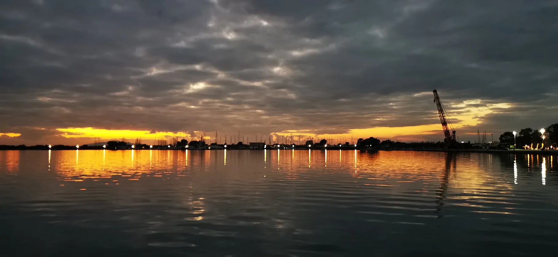 Messolonghi harbour at dusk under heavy clouds, lights reflected in calm water and the masts of the marina in the distance.