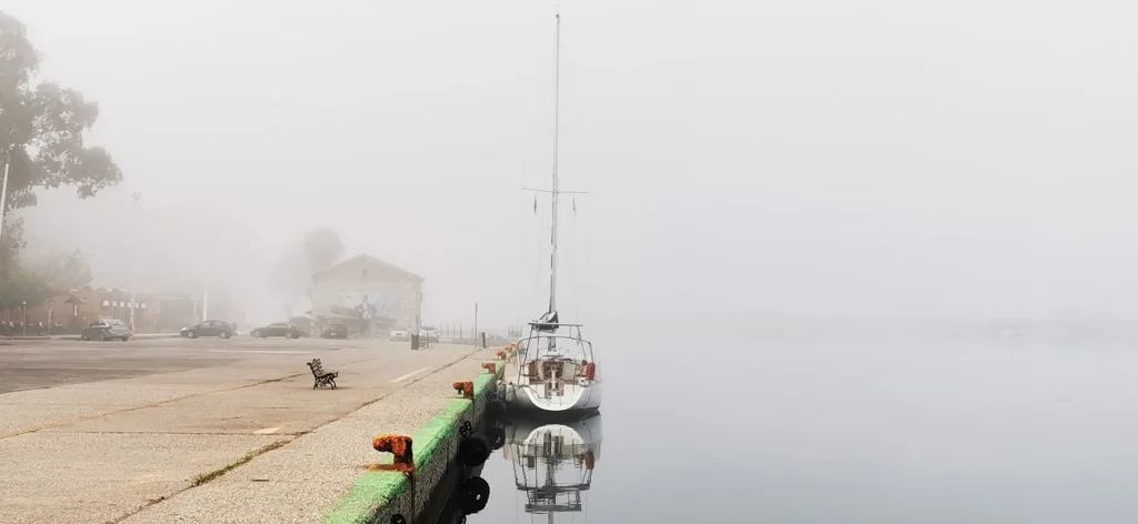 Water Mellon moored alone on the Messolonghi town quay on a foggy morning, her reflection in still water, rusty bollards and an empty bench on the quay.