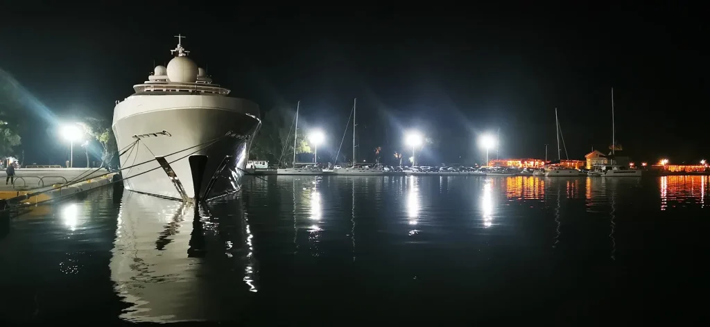 A large superyacht moored at the Messolonghi quay at night, its bow reflected in still water, with sailing boats and lights beyond.