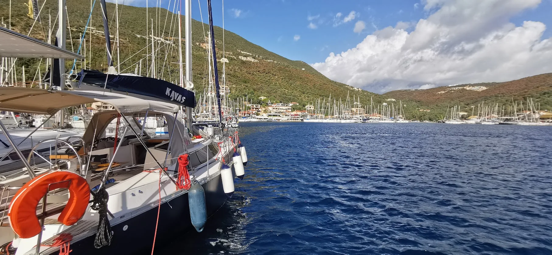 Water Mellon moored on a quay in a green Ionian bay, under a blue sky with white clouds.