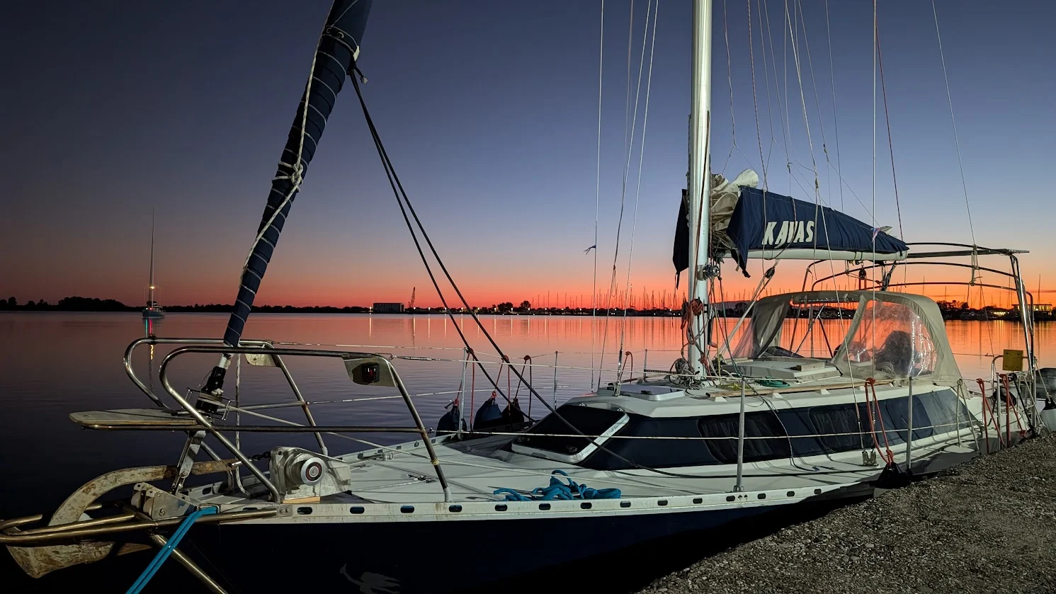 Water Mellon moored on a gravel shore at sunset, the sky deep blue above and orange on the horizon, calm water reflecting the light and the mermaid logo on her hull.